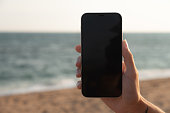 A girl holds a smartphone in her hand close-up with a black screen against the background of the sea. Technologies, modern methods of communication and remote work