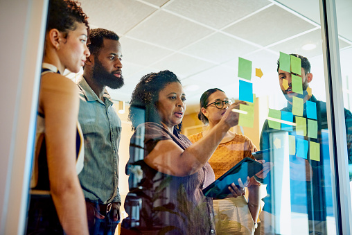 A group of diverse professionals are brainstorming ideas while standing around a glass wall with sticky notes.