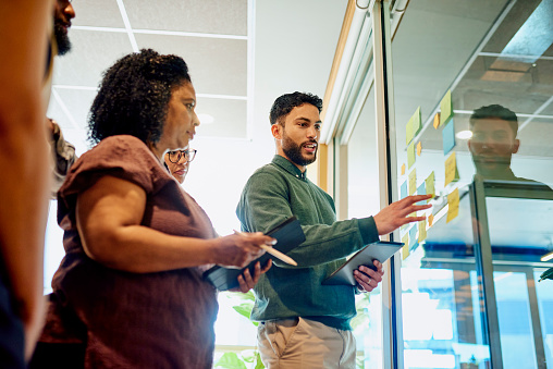 A group of diverse professionals brainstorm and collaborate, using sticky notes on a glass wall, in a modern office space.