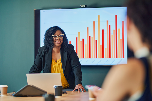 A successful businesswoman stands in front of a group of colleagues during a meeting. She is presenting financial data on a large 
screen and is smiling and looking at her colleagues.