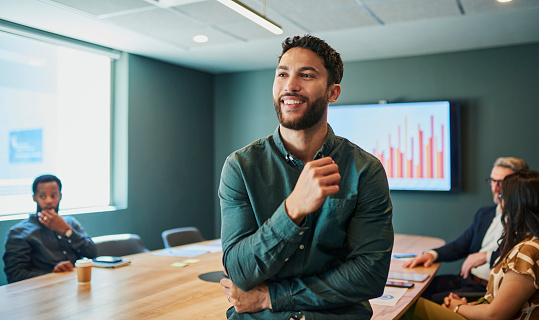 Confident young businessman standing in a modern office and smiling. He is wearing a casual shirt and has a beard. In the background, there are people sitting at a conference table.