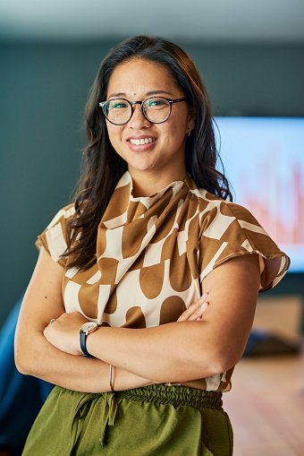 A young businesswoman with glasses is standing in an office, smiling confidently with her arms crossed.