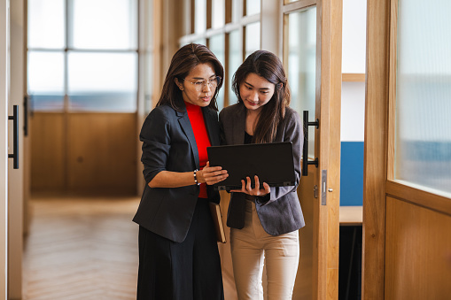 Two Asian Chinese female businesswomen collaborate, discussing ideas in a modern office corridor. The corporate professionals are engaged in deliberations regarding a new project and sharing innovative ideas in the workplace.