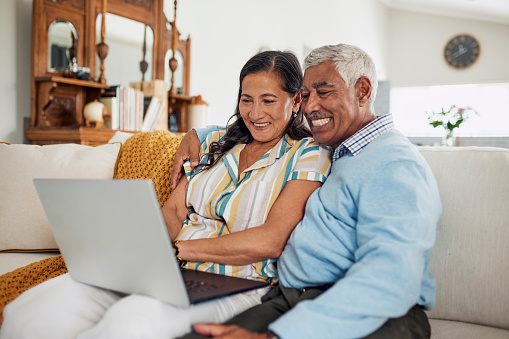 An elderly couple sits on a couch and smiles while looking at their laptop.