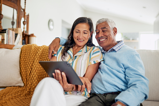 An elderly couple sitting on a couch and smiling while looking at a tablet.
