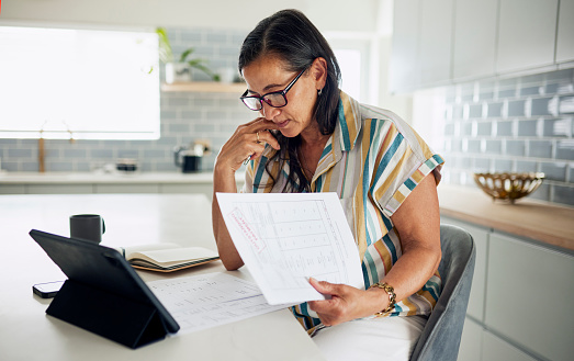A focused Latina woman in her 40s wearing glasses reads through important documents while sitting at her kitchen counter. She has a tablet on the counter and is holding her chin in thought.