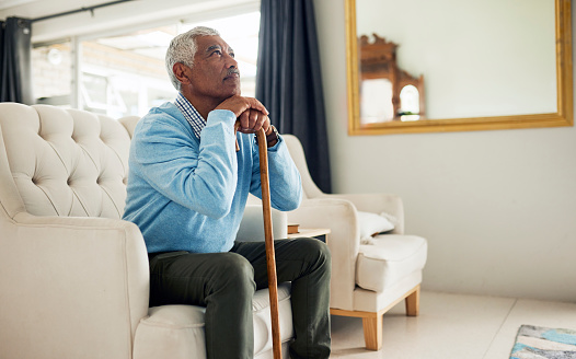 An elderly man sits on a couch, leaning on a cane and looking lost in thought. He is wearing a blue sweater and khaki pants, and the room is furnished with white furniture and a mirror.