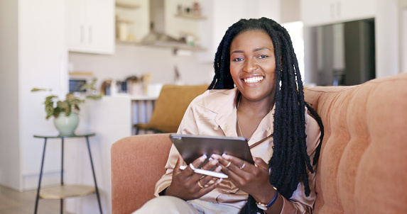A young black woman is sitting on a couch in her home, smiling while using a tablet. She is wearing a casual outfit and has her hair in long braids.