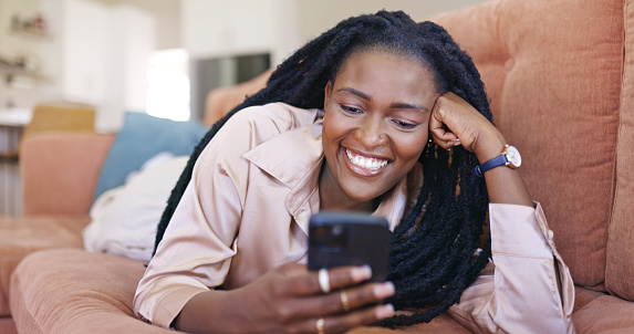 A young woman is lying on a couch and smiling while texting on her phone. She is wearing a pink silk shirt and has her hair in long braids.