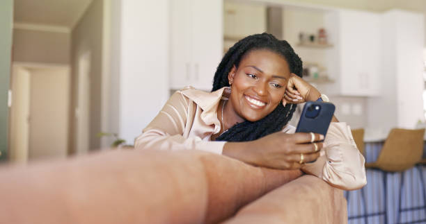 istock Beautiful black woman smiling while relaxing on couch and using smartphone. 1887470298