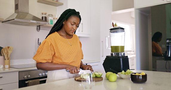 A young African American woman is making a healthy green smoothie in her kitchen. She is cutting up apples and putting them in the blender.