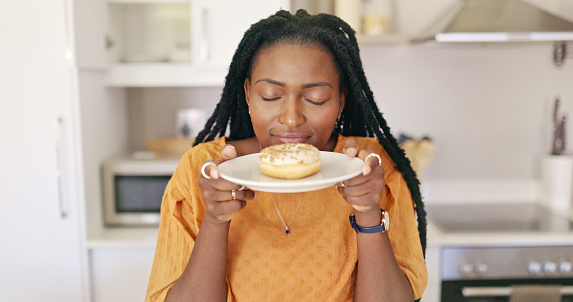 A young African American woman closes her eyes and inhales the sweet scent of a donut held on a plate in her hands.