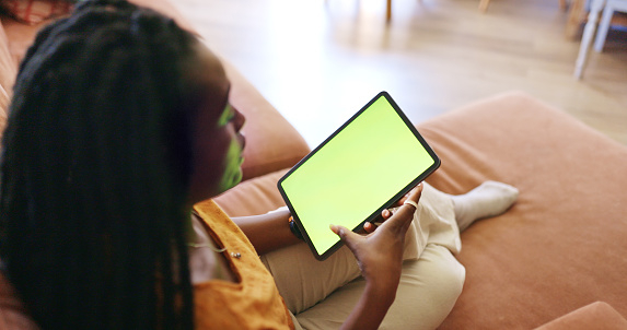 A young woman sits on a couch in her home and uses a tablet. She is swiping the screen with her finger