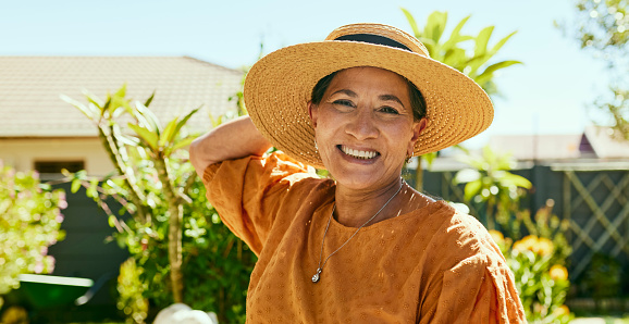 A happy looking elderly woman wearing a wide-brimmed straw hat is gardening in her backyard on a sunny day