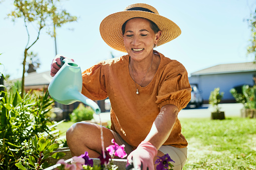 A smiling woman wearing a hat is gardening and watering her plants.