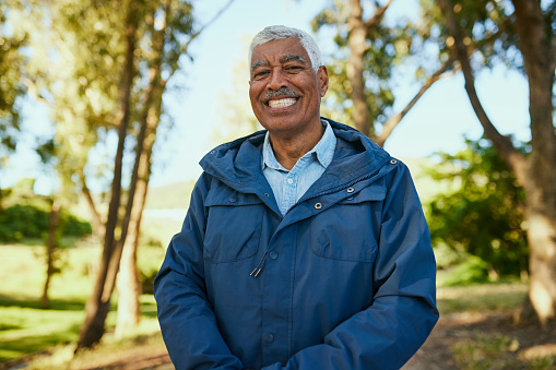 A cheerful elderly man with gray hair and a mustache is captured smiling outdoors. He is wearing a blue windbreaker jacket and a light blue shirt. The background showcases a blurred park scene with trees and sunlight filtering through.