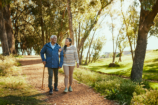 An elderly couple smiles and walks arm in arm through a park, surrounded by lush trees and bathed in warm sunlight.