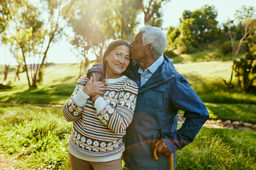 An elderly couple is lovingly embracing each other outdoors in a natural setting, with the man kissing the woman on the cheek