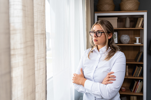 Depressed woman standing with arms crossed looking through window at home. Female issue mental health behavior sad person disappointed in life after relationship breakup or divorce. Depression concept