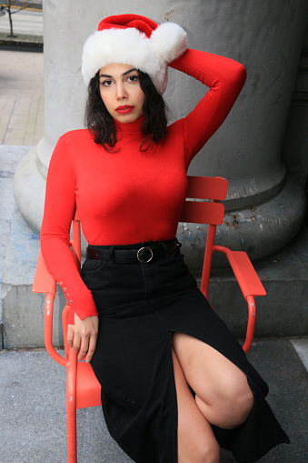 An Iranian model sitting in a red chair. She is wearing a Santa hat, medium length black hair, makeup, a red turtleneck, and a  black skirt.