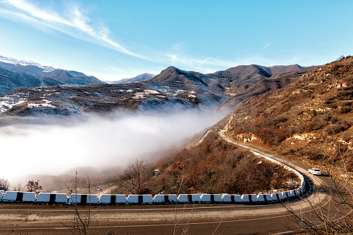 A road in the mountains of Dagestan with steep descents and turns, passing along the edge of a deep gorge. There is a thick white fog in the gorge. There is snow melting in the sun on the mountains.