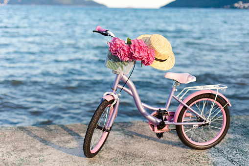 Female romantic bicycle with pink hydrangea flowers and straw hat on sea beach. Concept of children riding on coastline, tenderness, childhood, summer.