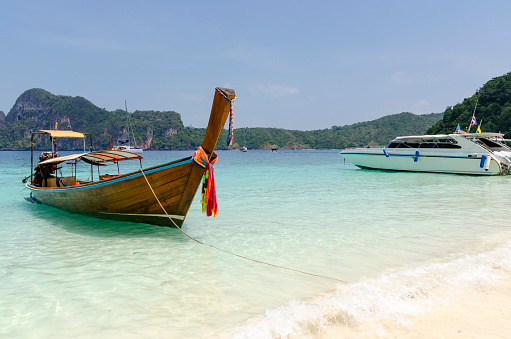 A vibrant image of the Phi Phi Islands, Thailand, where the bustling tourism is matched by the stunning natural scenery. Traditional long-tail boats and sleek power boats navigate the azure waters, framed by towering limestone cliffs and lush greenery. This landscape is a paradise for visitors, offering a striking combination of human activity and breathtaking tropical beauty.