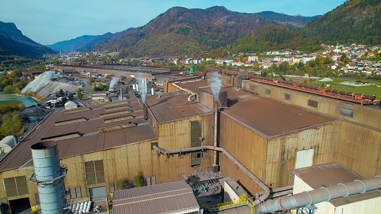 AERIAL: Factory surrounded with alpine landscape in vibrant colors of fall season. Industrial infrastructure located in picturesque valley surrounded city and mountains glowing in vivid autumn colors.