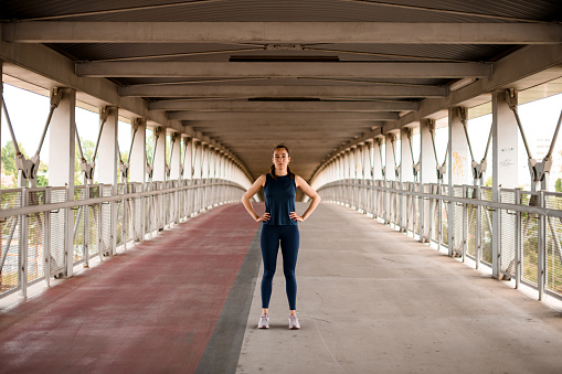 Outdoor shot of sporty woman dressed in activewear looking into camera on bridge , blurred background. Full length photo. Healthy lifestyle.