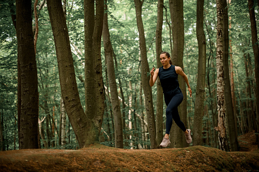 Woman with long dark hair in pony tail running on hilly forest area between tall trees. Sport and healthy lifestyle concept.