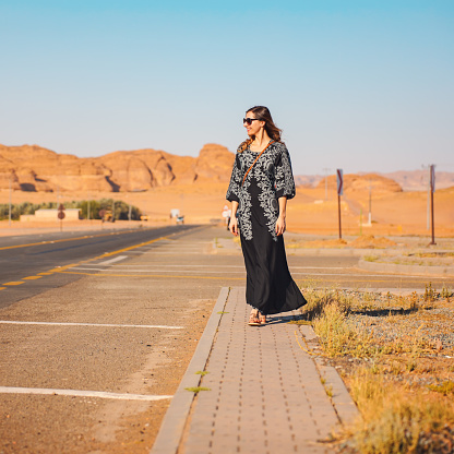Young woman in long black dress walking on pavement near road - desert rocky landscape, typical for AlUla, in background