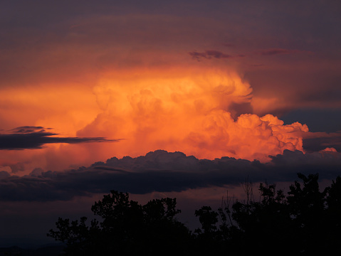 Dramatic cumulonimbus cloud rises above treetops and glows in sunset colours. Magnificent view of summer evening thunderstorm in vibrant colours. Storm clouds forming on summer sky over countryside.