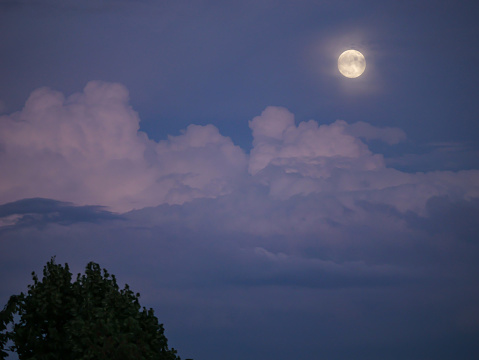 Beautiful mystic full moon rising above stormy clouds in bluish hues of twilight. Amazing colors of blue hour after summer sunset. Magnificent view of mystic moon rising between moonlit stormy clouds.