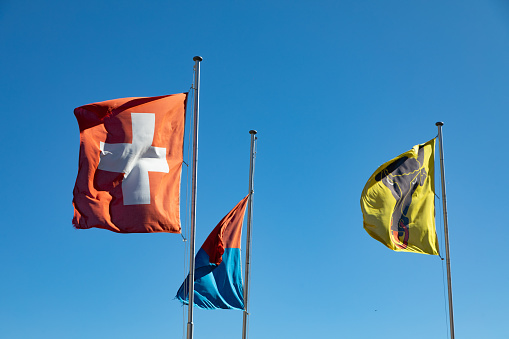 Swiss flag with Uri Canton and Ticino Canton flags against a blue sky at Saint Gotthard Pass, Switzerland