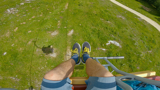 POV: Spring chairlift ride with view of male feet above green mountain pastures. Adventurous panoramic ride with ski lift on a sunny spring day passing by beautiful spring-flowering alpine meadows.