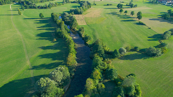 AERIAL: Meandering river, surrounded by lush greenery, winds through the fields. Picturesque countryside with slow-flowing river and vibrant green meadows in flattering summer afternoon sunlight.