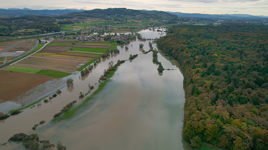 AERIAL: Flood water from muddy swollen river spilling over the rural landscape. Flooded countryside after abundance of rain in autumn. Rising water spreading over grassy riverside, fields and woods.
