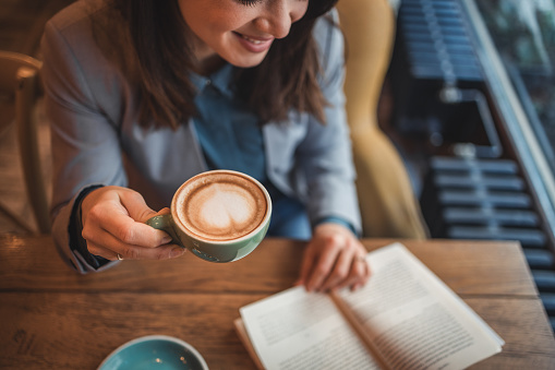 Young woman elegantly dressed, for work, drinking coffee and reading a book in a cafe, enjoying the morning and morning routine, having a break at work and using it to relax