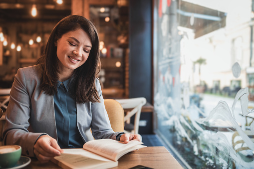 Young woman elegantly dressed, for work, drinking coffee and reading a book in a cafe, enjoying the morning and morning routine, having a break at work and using it to relax