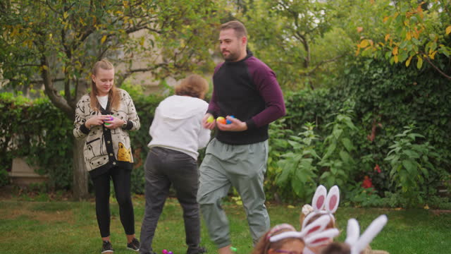 Adults hiding Easter eggs in background while excited children sitting on grass and waiting with their eyes closed in back yard garden