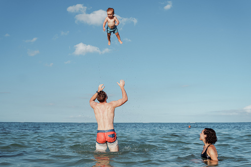 A Caucasian man who is at the beach in Hawaii with his family, playfully throws his Eurasian toddler son up into the air while his wife swims nearby and watches.