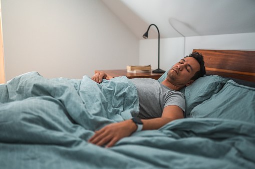 3/4 length image of a handsome, biracial man sleeping in bed at dawn, wearing a wrist watch. Lying down, eyes closed, hand stretched out on the bed.