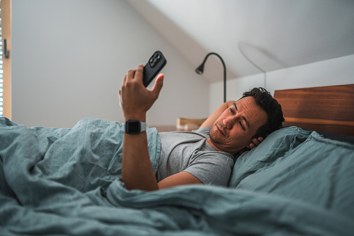 Waist up image of a mid adult, Asian man reading mail on his smart phone while still in bed, looking sleepy , covered with a blanket over his stomach. Lying down, one hand  behind his head. Shot over the other side of bed.