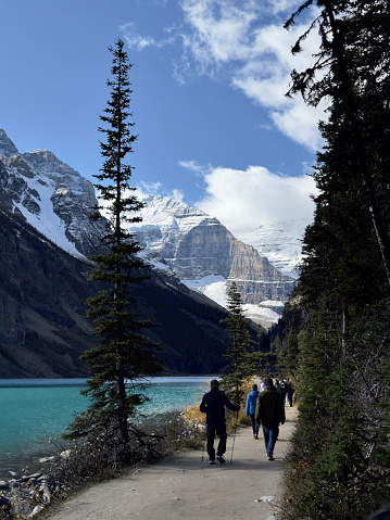 Medium shot of hikers on a trail winding its way around Lake Louise, framed by its majestic snowcapped Mount Lefroy and others