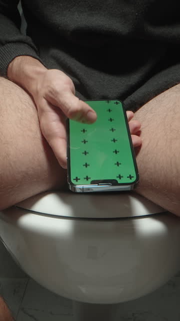 Vertical video. Chroma Key Layout on a Smartphone in the Hand of a Man Sitting on a Toilet, Low Angle View, Dolly Shot. Concept of Scrolling Through a Social Media Feed or Online Store.