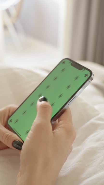 Vertical video. Smartphone with a Green Screen Mockup and Tracking Markers, a woman scrolling through a social media feed while sitting on a white bed.