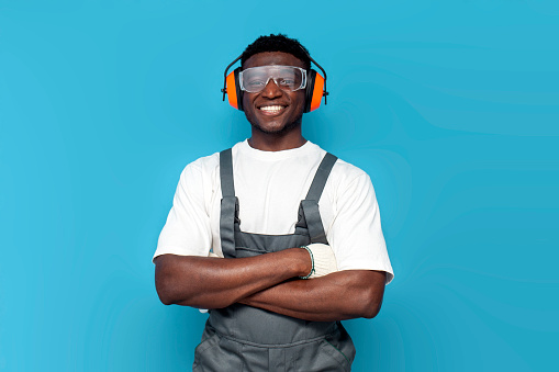 african american male builder worker in uniform stands with his arms crossed on blue isolated background, handyman in overalls and goggles with construction headphones