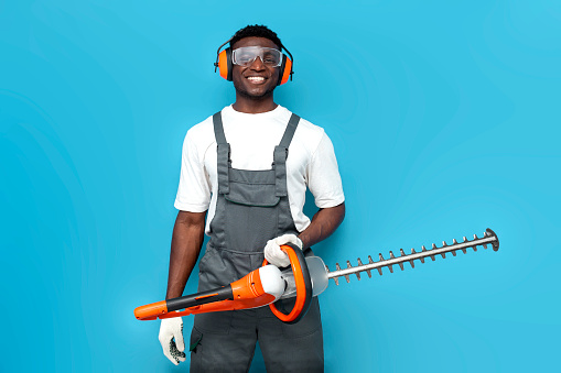 garden worker in uniform holding electric brush cutter on blue isolated background, african american man in overalls goggles and construction headphones with gardening tool
