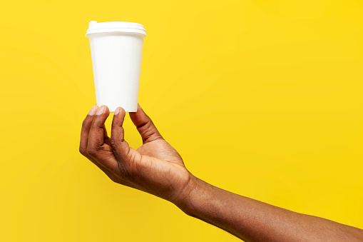 african american man's hand holds white paper cup of coffee on yellow isolated background, hand offers and advertises drink with mock-up, close-up
