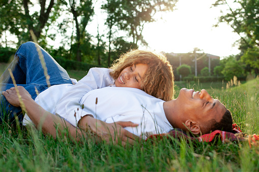 african american romantic young couple lies on the grass in the park and sleeps in nature, happy family rests and dreams outdoors, woman hugs her boyfriend and smiles